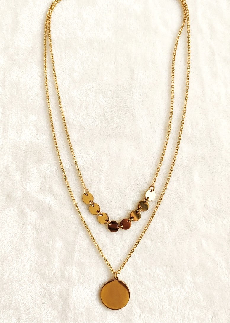 A double-layered anti tarnish gold necklace featuring a delicate chain with small metallic disc charms and a larger flat round pendant, displayed on a textured white fabric background.