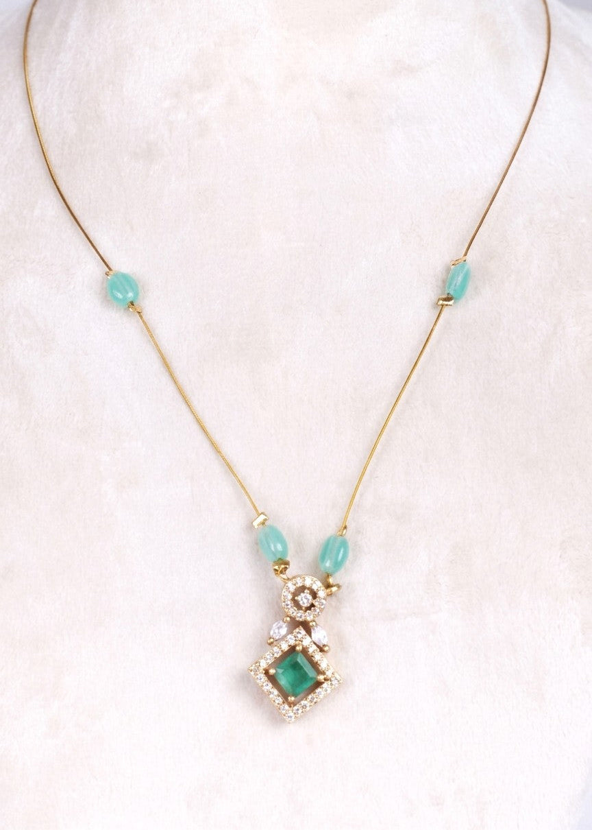 A gold-tone necklace featuring a central pendant with a green square gemstone surrounded by clear crystals, accented by three light blue beads on the chain, displayed on a soft white fabric background.