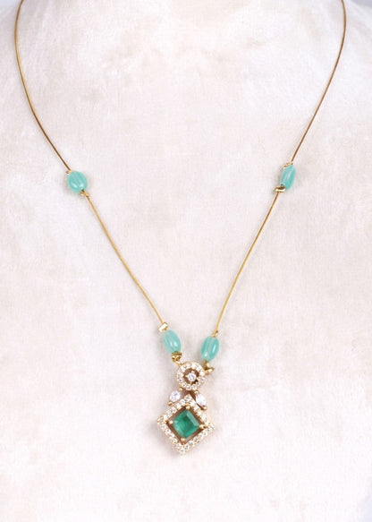 A gold-tone necklace featuring a central pendant with a green square gemstone surrounded by clear crystals, accented by three light blue beads on the chain, displayed on a soft white fabric background.