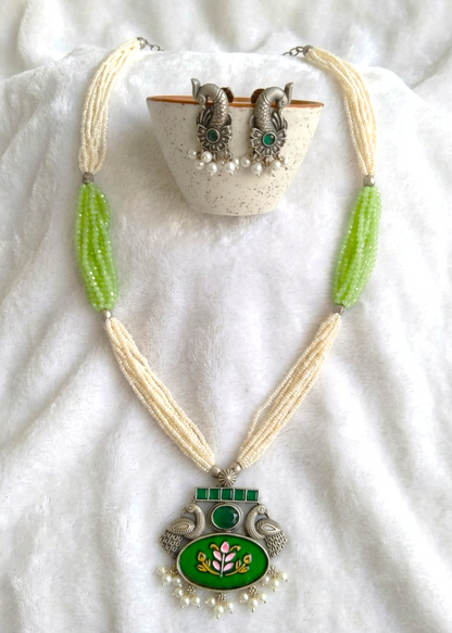 A traditional Indian jewelry set featuring a necklace and matching earrings. The necklace has cream-colored multi-strand beads, bright green bead accents, and a silver-plated pendant with green enamel, a central green and gold floral motif, and small pearl-like beads at the bottom.