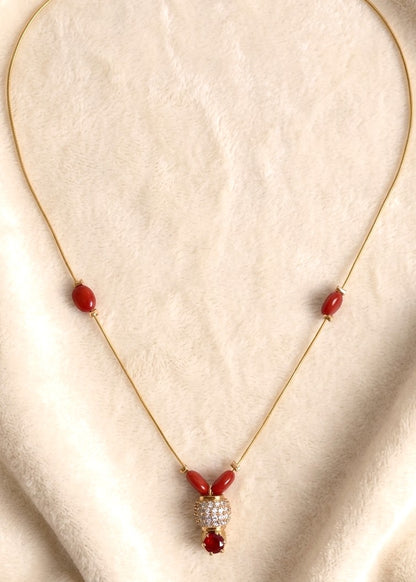 A gold chain necklace with red bead accents and a central pendant featuring a clear crystal-encrusted heart and a red jewel, displayed on a textured beige fabric background.