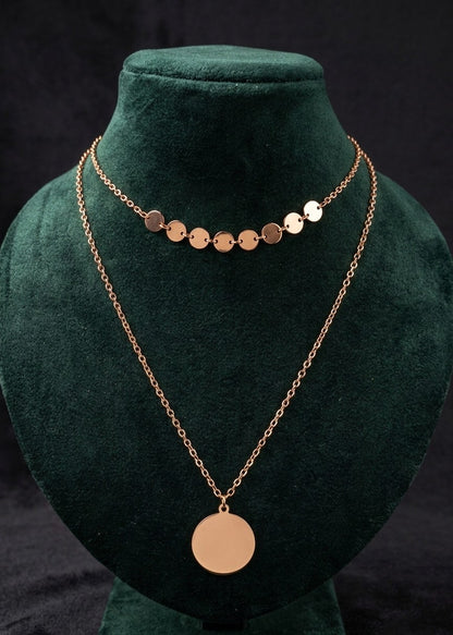 A rose gold double-layer necklace displayed on a dark green velvet jewelry bust. The necklace features a short chain with multiple small flat circular disc charms and a longer chain with a larger round disc pendant.