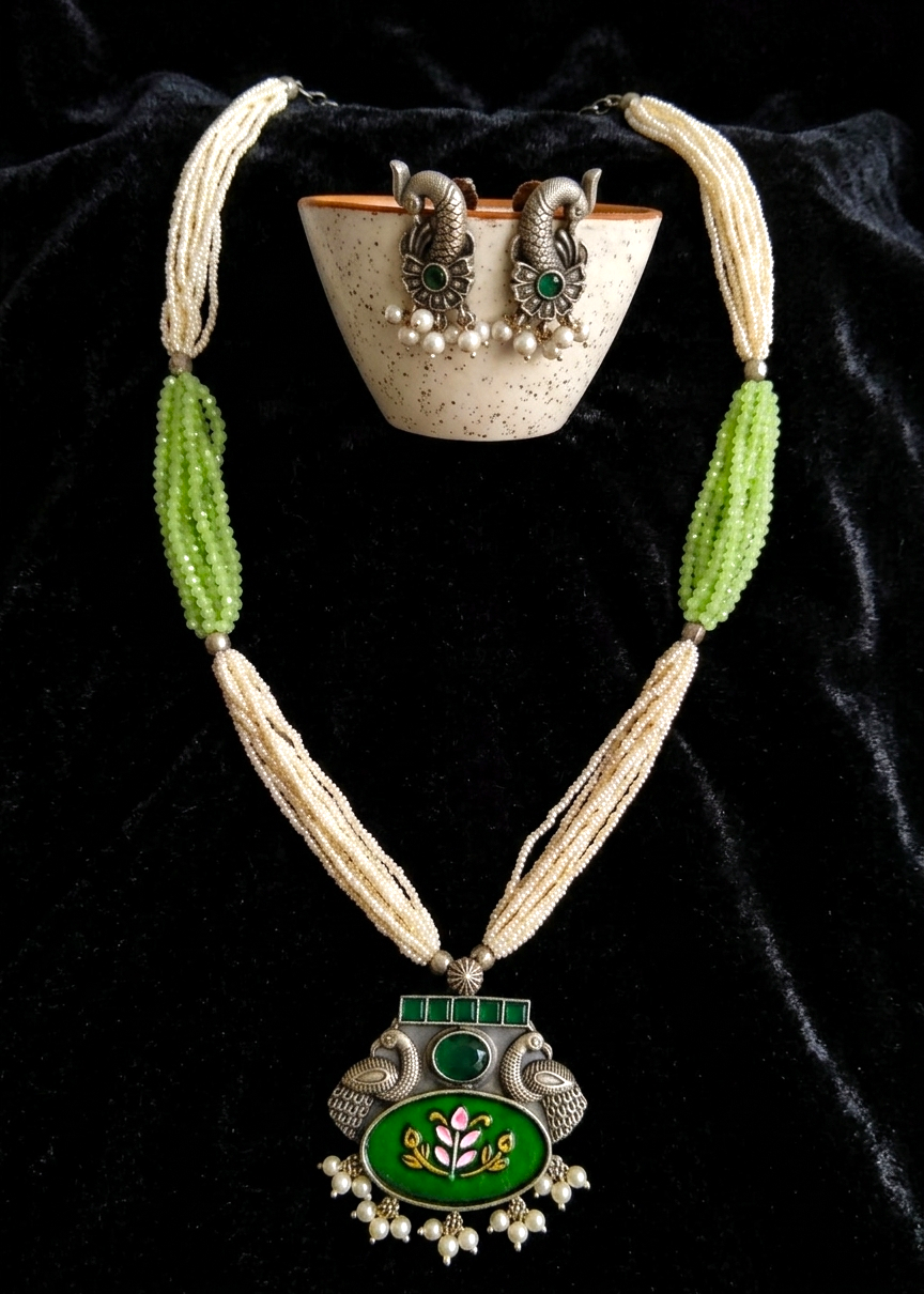 A traditional Indian jewelry set displayed on a black velvet background, featuring a long beaded necklace with a green and silver pendant, a matching bowl, and earrings.