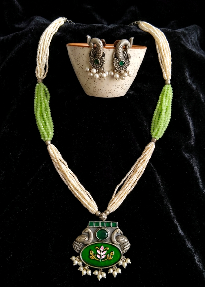 A traditional Indian jewelry set displayed on a black velvet background, featuring a long beaded necklace with a green and silver pendant, a matching bowl, and earrings.