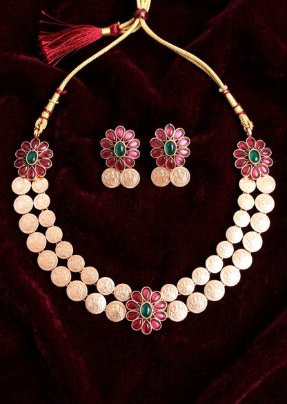 A gold-toned jewelry set featuring a coin-studded necklace and matching earrings, decorated with pink flower petals and green stone centers, displayed on a dark red velvet background.