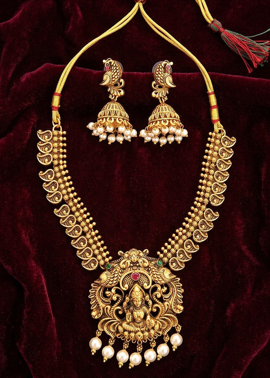 Gold-toned traditional necklace and matching earrings displayed on a deep burgundy velvet background. The necklace features an ornate Lakshmi pendant, beaded strands, and dangling pearl-like beads, with matching earrings placed above it.