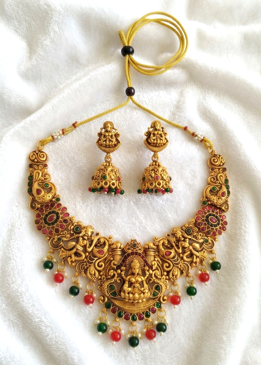 An ornate gold-toned jewelry set featuring a necklace and matching earrings with detailed filigree, a central deity motif, and red and green bead accents, displayed on a white textured fabric background.