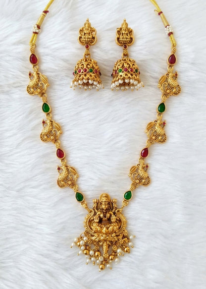 Gold-toned traditional Indian jewelry set featuring a Lakshmi pendant necklace, matching dangle earrings, and decorative chain links with red and green stone accents, displayed on a white faux fur background.