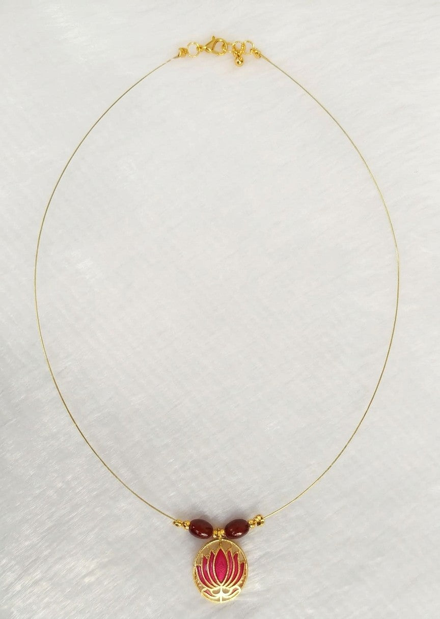 A delicate gold chain necklace featuring a central pendant with two small dark red beads and a gold lotus flower enclosing a pink enamel center, displayed on a textured white background.