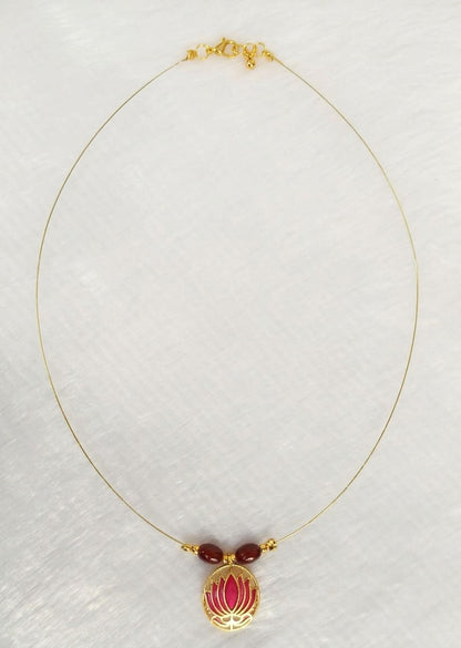 A delicate gold chain necklace featuring a central pendant with two small dark red beads and a gold lotus flower enclosing a pink enamel center, displayed on a textured white background.