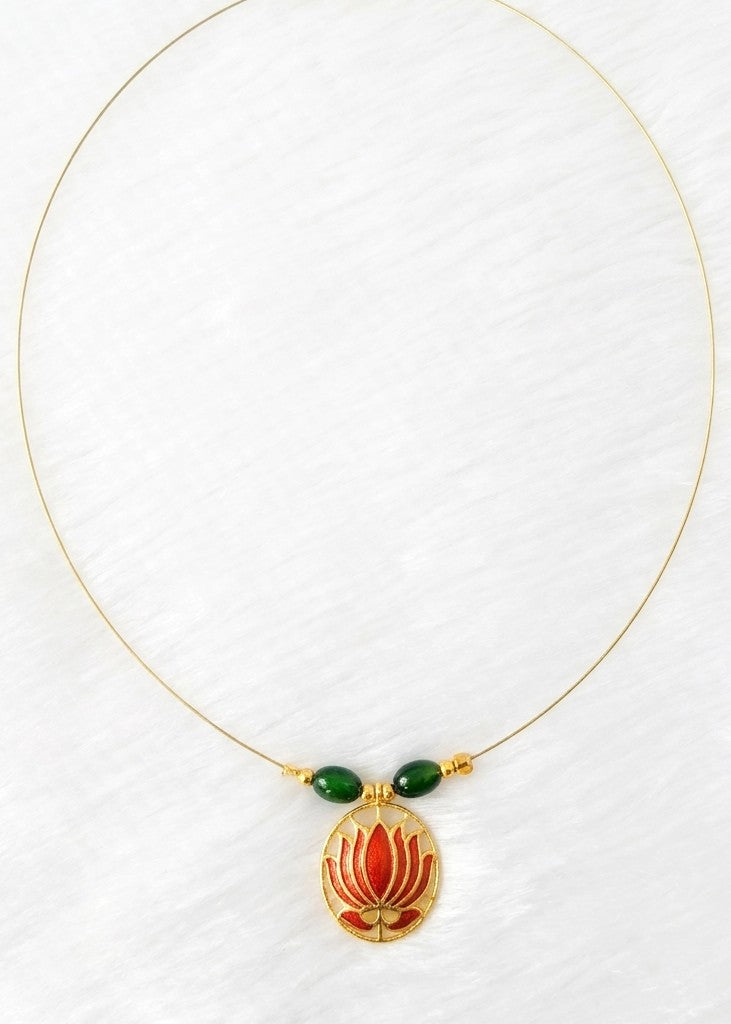 A delicate gold necklace featuring a central gold pendant with red and orange floral detailing, accented by two small green beads on the chain, displayed on a textured white background.