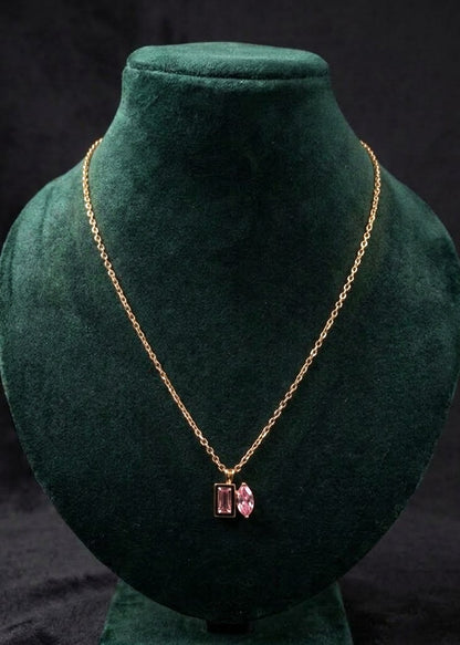 A gold chain necklace with a rectangular pink gemstone pendant, displayed on a dark green velvet jewelry bust against a black background.