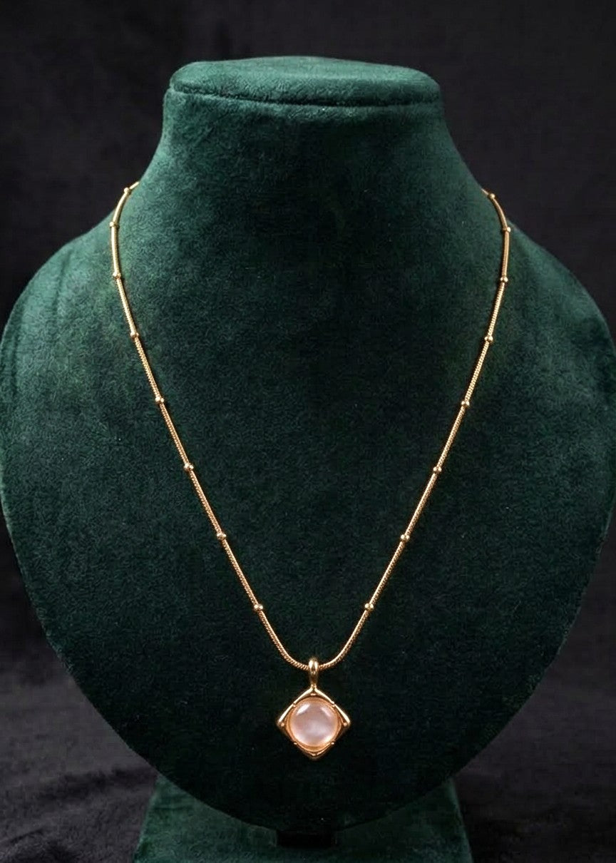 A gold beaded chain necklace with a diamond-shaped pendant featuring a pale pink stone, displayed on a dark green velvet jewelry bust against a black background.