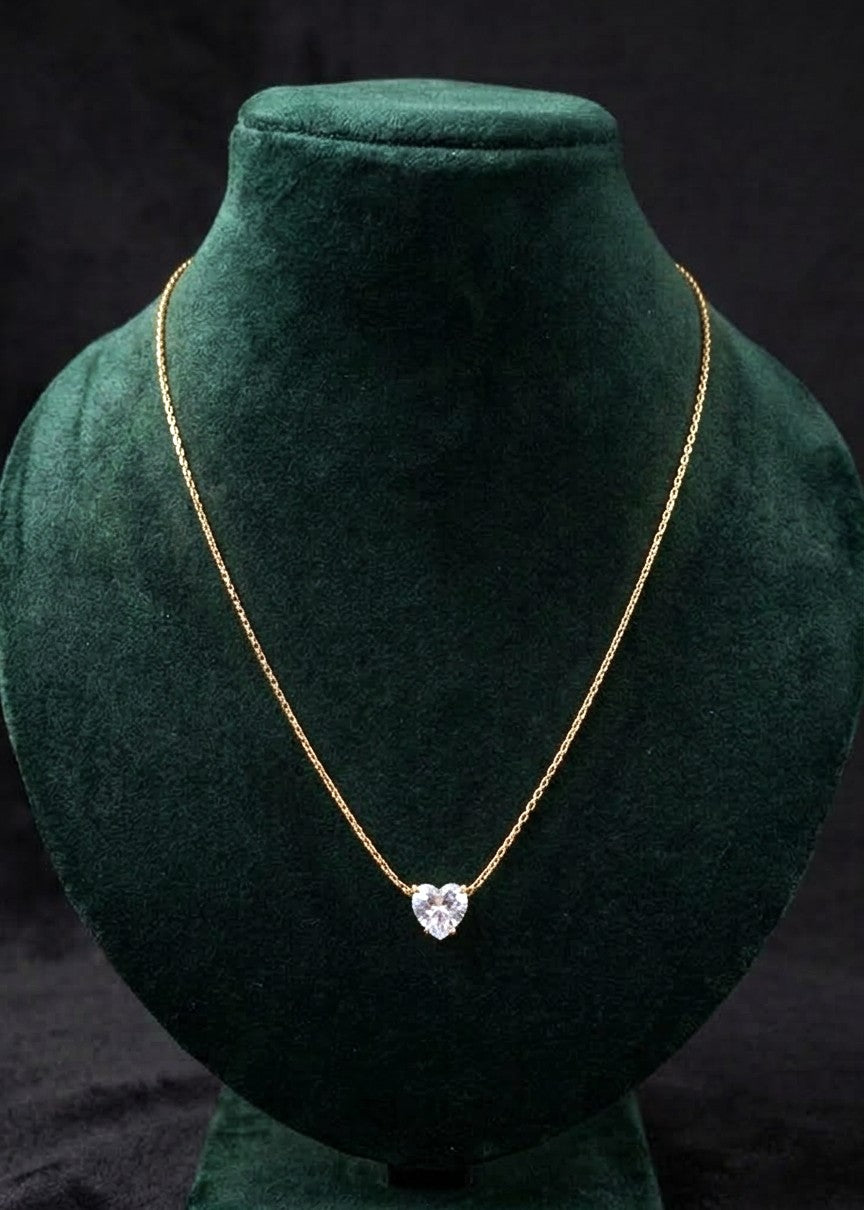 A delicate gold chain necklace with a heart-shaped clear gemstone pendant, displayed on a dark green velvet jewelry bust against a black background.