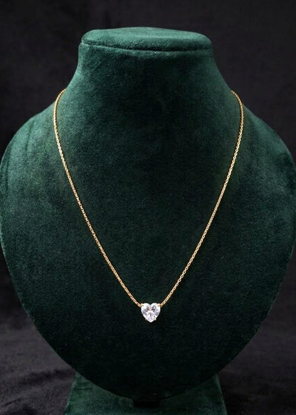 A delicate gold chain necklace with a heart-shaped clear gemstone pendant, displayed on a dark green velvet jewelry bust against a black background.