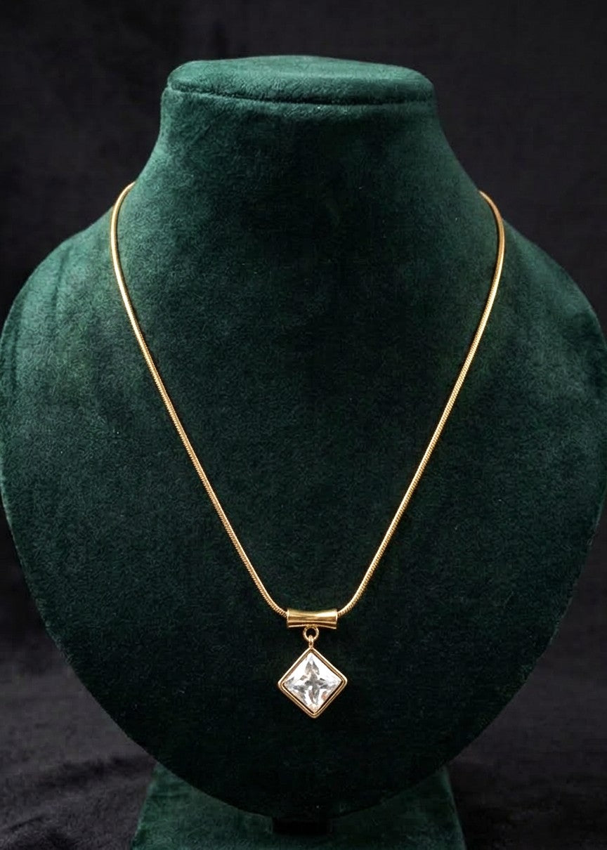 A gold chain necklace with a diamond-shaped pendant featuring, displayed on a dark green velvet jewelry bust against a black background.