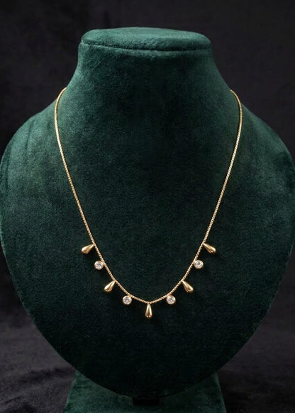 A delicate gold chain necklace with small teardrop-shaped gold charms and clear round stones, displayed on a dark green velvet jewelry bust against a black background.