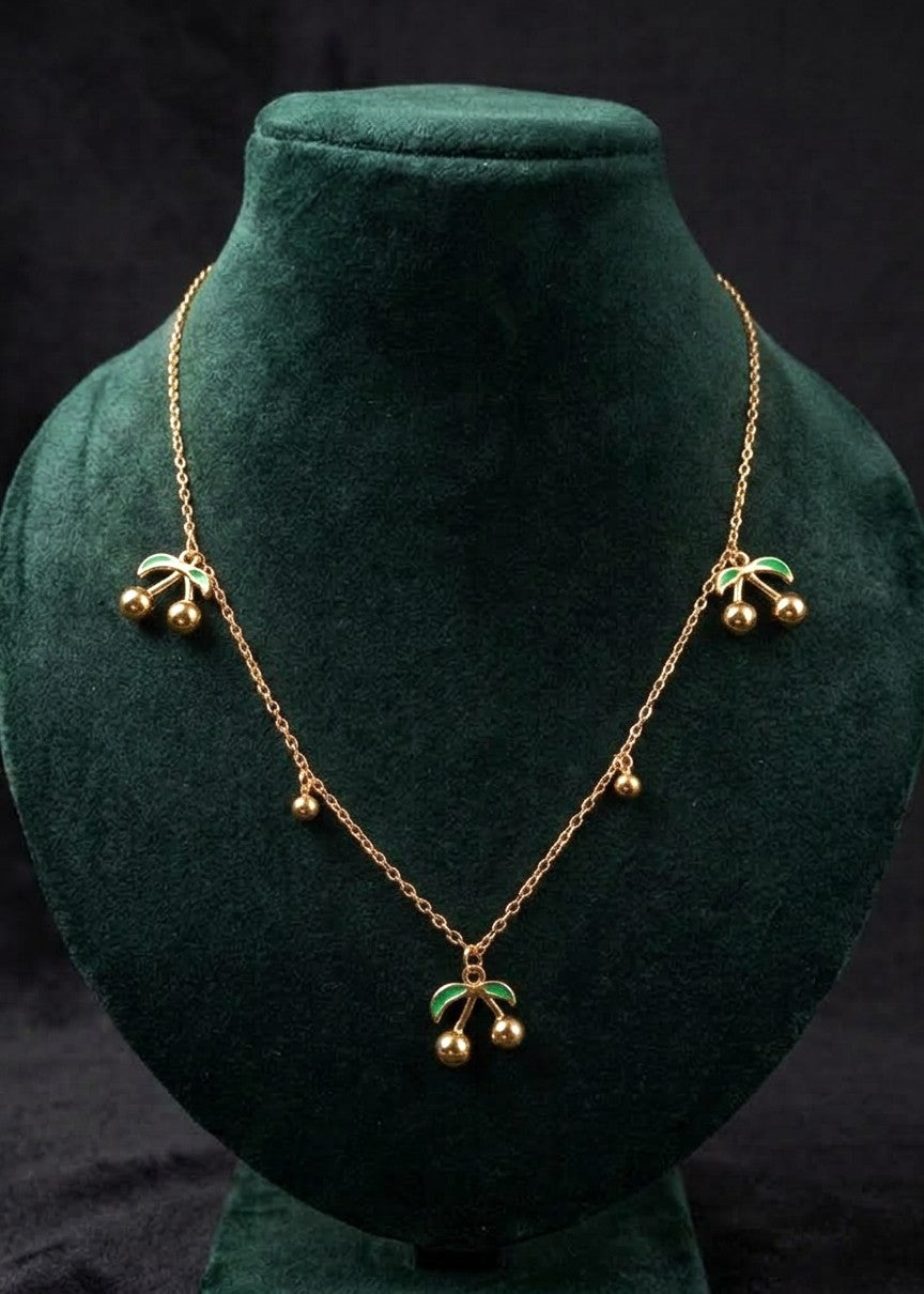 Celine gold-toned necklace with three cherry charms, green enamel leaves and dangling beads on a dark green display bust.