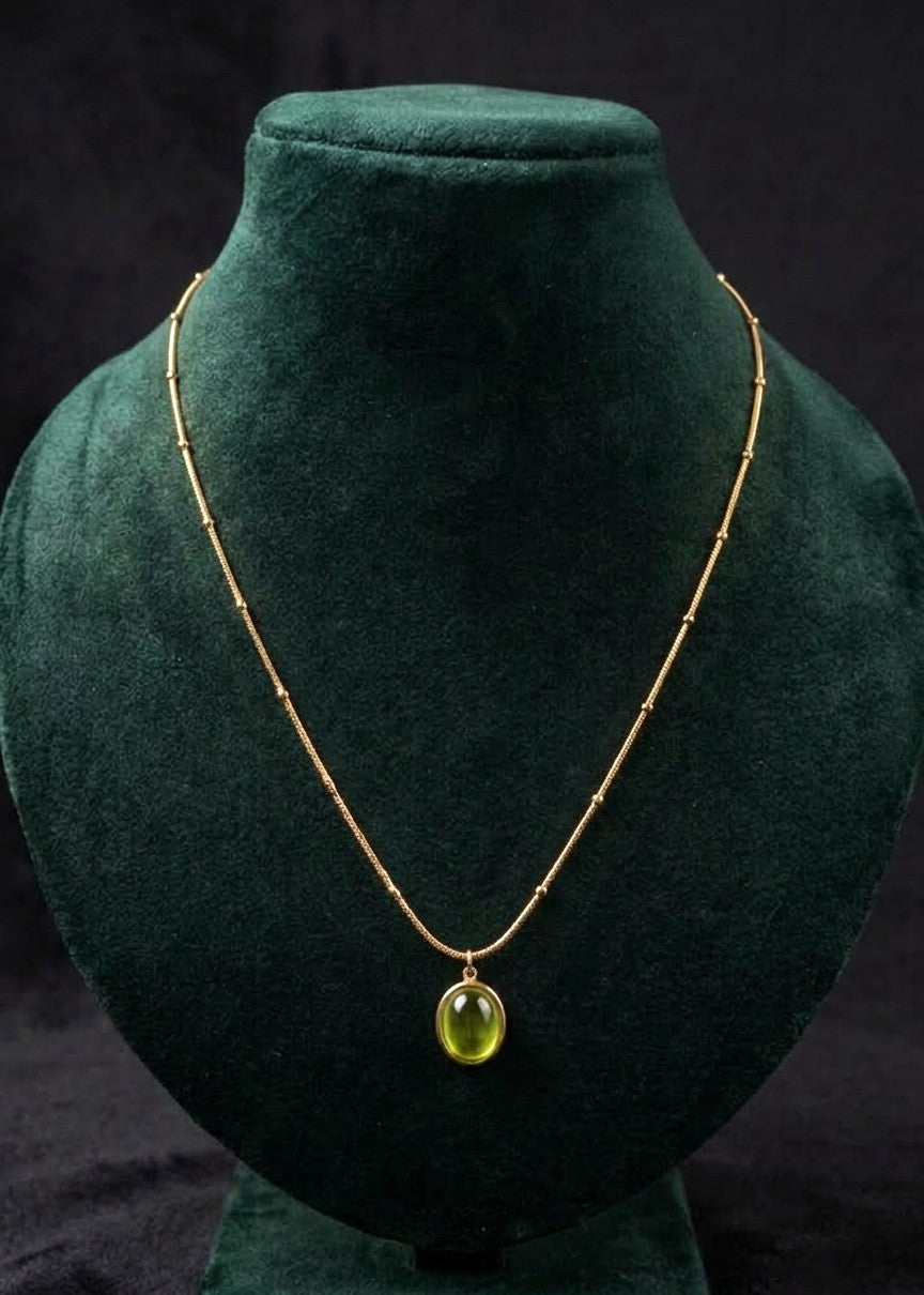 A gold chain necklace with a small oval green pendant, displayed on a dark green velvet jewelry bust against a black background.