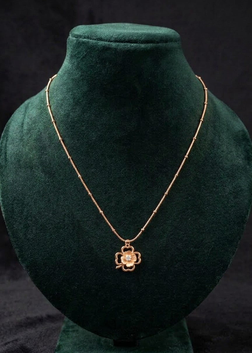 Mahnoor gold-toned clover pendant necklace with a beaded chain and white crystal centre on a dark green display bust.