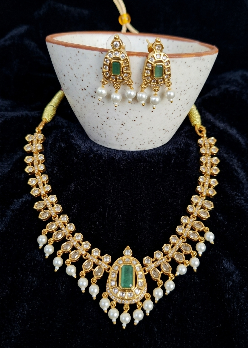 Gold-plated Indian Kundan jewelry set featuring a necklace and matching earrings with green stone accents and white pearl drops, displayed on a speckled white ceramic stand against a black velvet background.