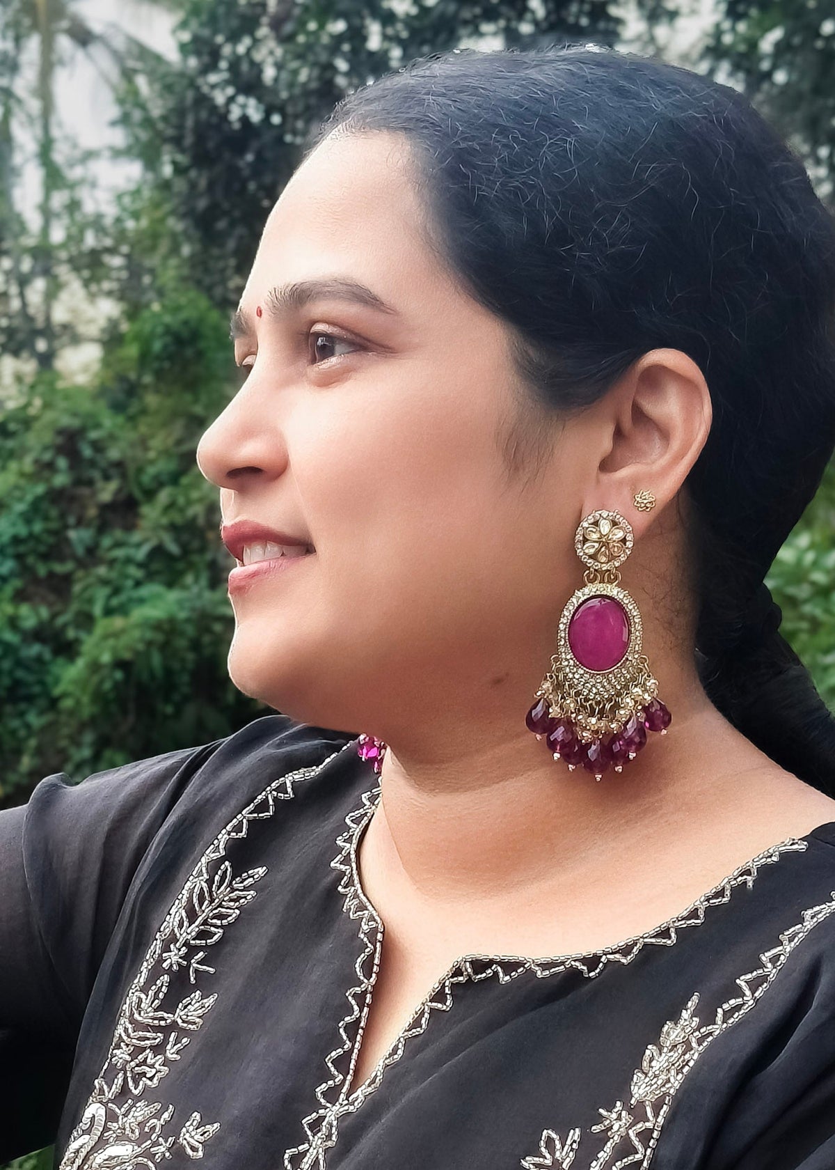 A woman in profile wearing ornate gold-toned dangle earrings with a prominent magenta-pink stone and purple bead fringe.