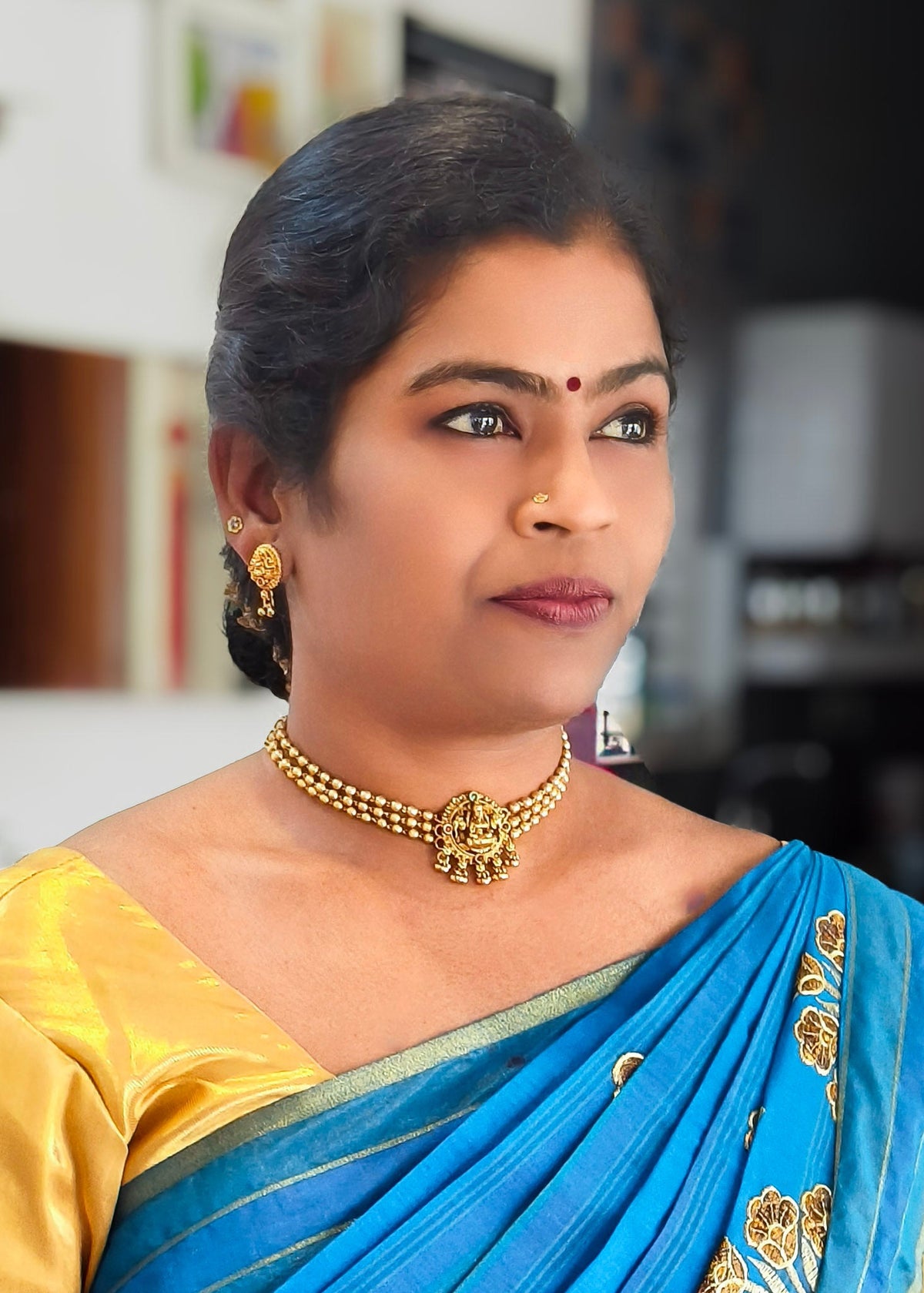 A woman wearing a bright blue and yellow sari with gold jewelry, photographed indoors with a softly blurred background.