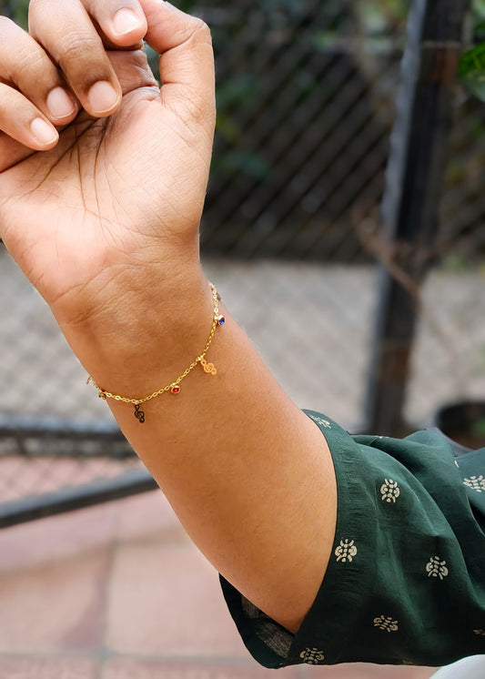 A close-up of a person's wrist wearing a delicate gold chain bracelet with small dangling charms, including a butterfly and a small red heart, against a blurred outdoor background.