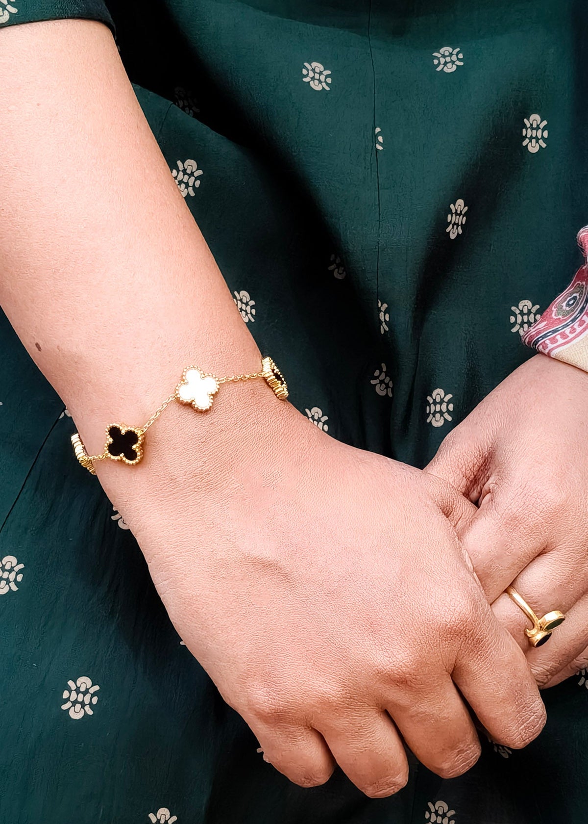A gold chain bracelet with three clover-shaped charms, two black and one white, worn on a person's wrist. The person is wearing a dark green printed dress and a gold ring on their finger.