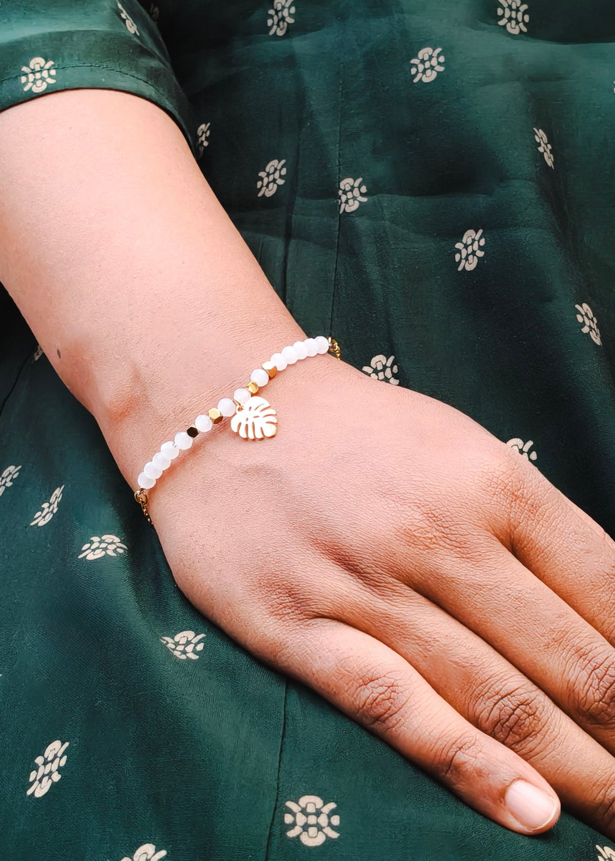 A close-up of a person's hand and wrist wearing a delicate gold bracelet adorned with white beads and a small gold leaf charm, set against a dark green patterned fabric.