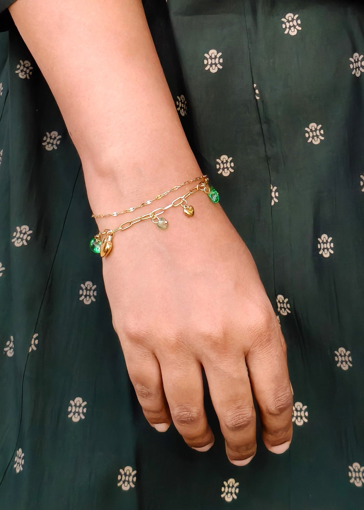 A close-up of a person's hand and wrist wearing a delicate double-strand gold bracelet with small green and white bead details and tiny charms, set against a dark green patterned dress.