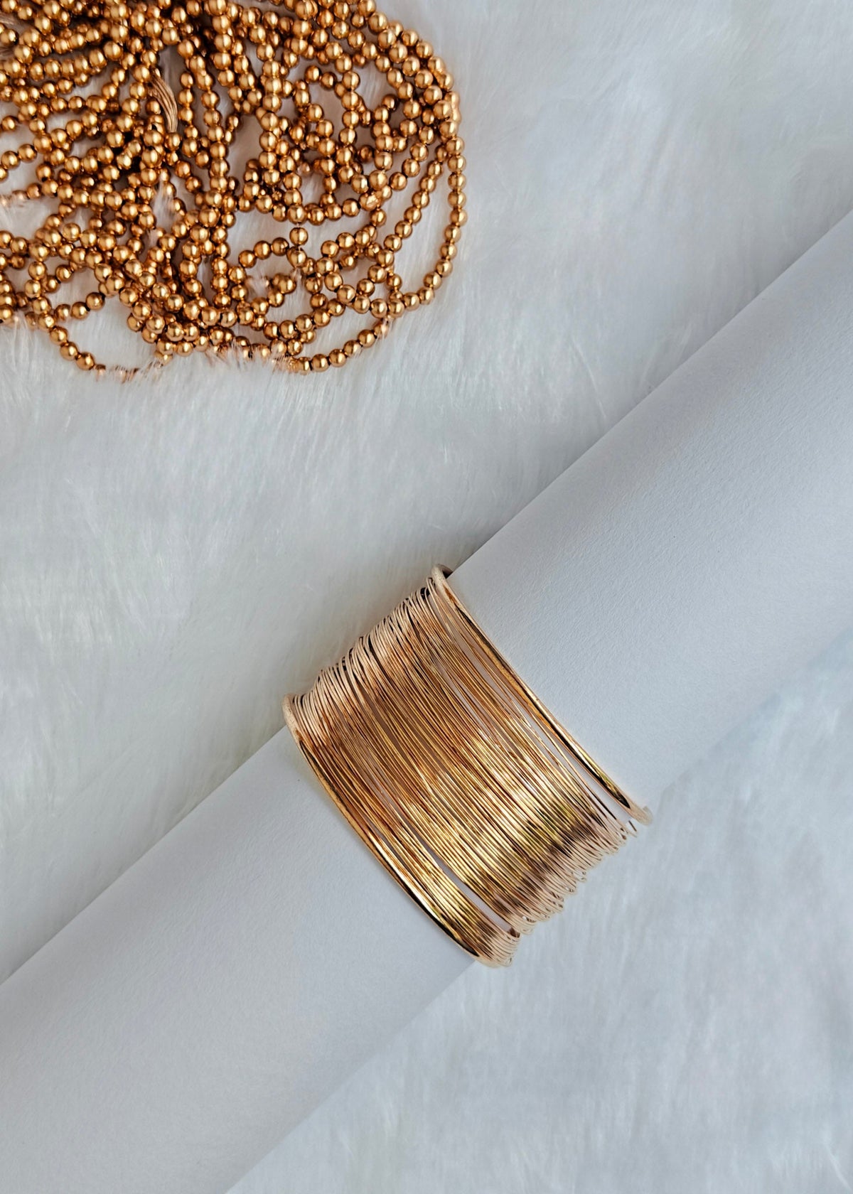A wide, multi-strand gold wire cuff bracelet displayed on a white cylindrical stand, with a matching gold beaded necklace resting on a white faux fur surface in the upper left.