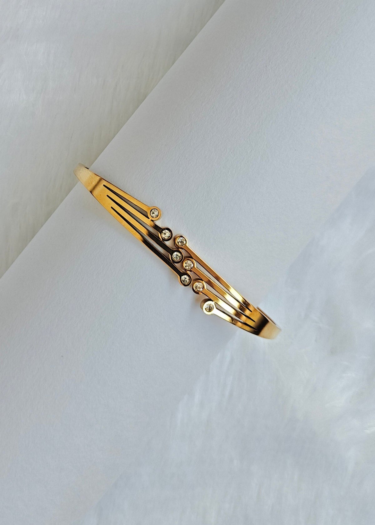 A gold-toned bangle bracelet with a geometric open design and small clear stones, displayed on a soft white textured surface.
