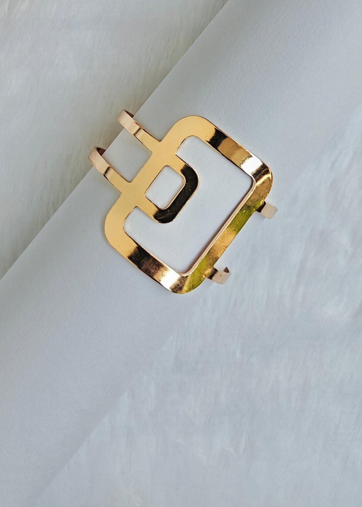A gold-toned metal cuff bracelet with a square openwork design and a white inlay, displayed on a soft white textured surface.