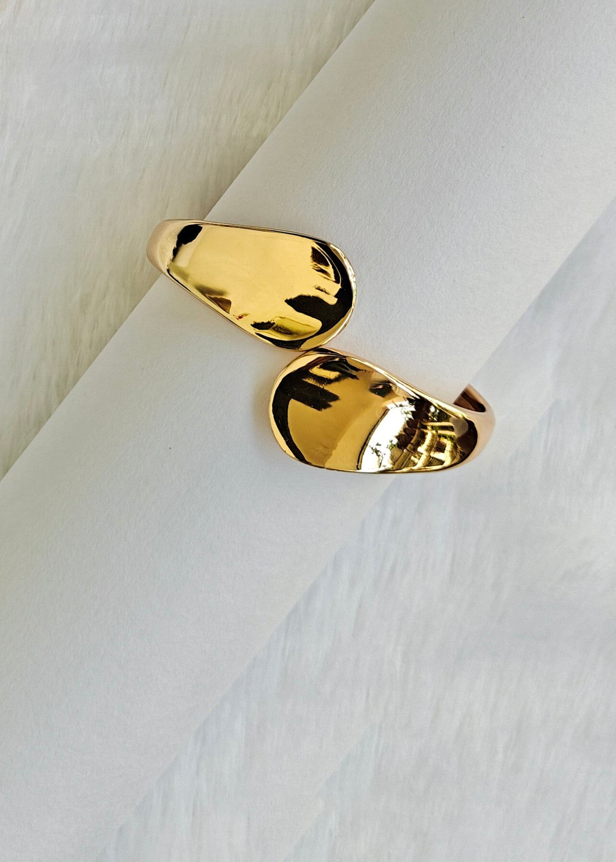 A pair of gold-toned abstract capsule-shaped rings displayed on a white cylindrical stand against a soft white textured background.