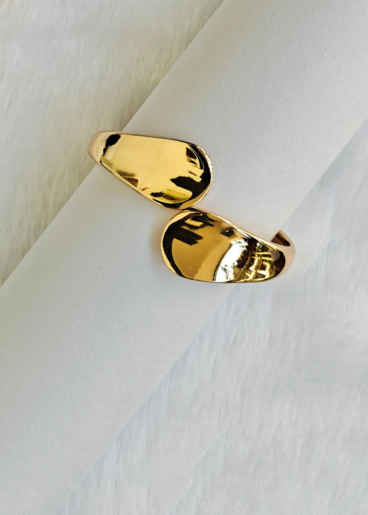 A pair of gold-toned abstract capsule-shaped rings displayed on a white cylindrical stand against a soft white textured background.