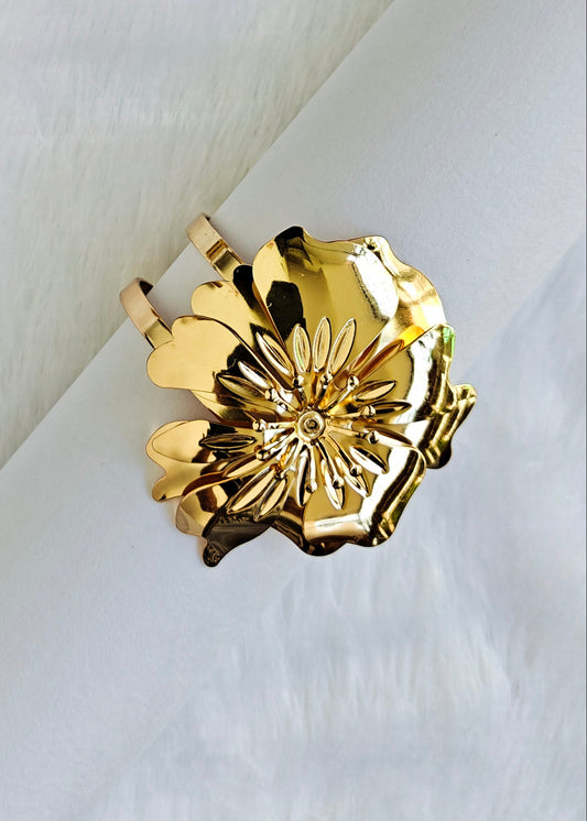 A gold-toned metal ring featuring a sculpted flower design with layered petals and a central sunburst motif, displayed on a textured white surface.