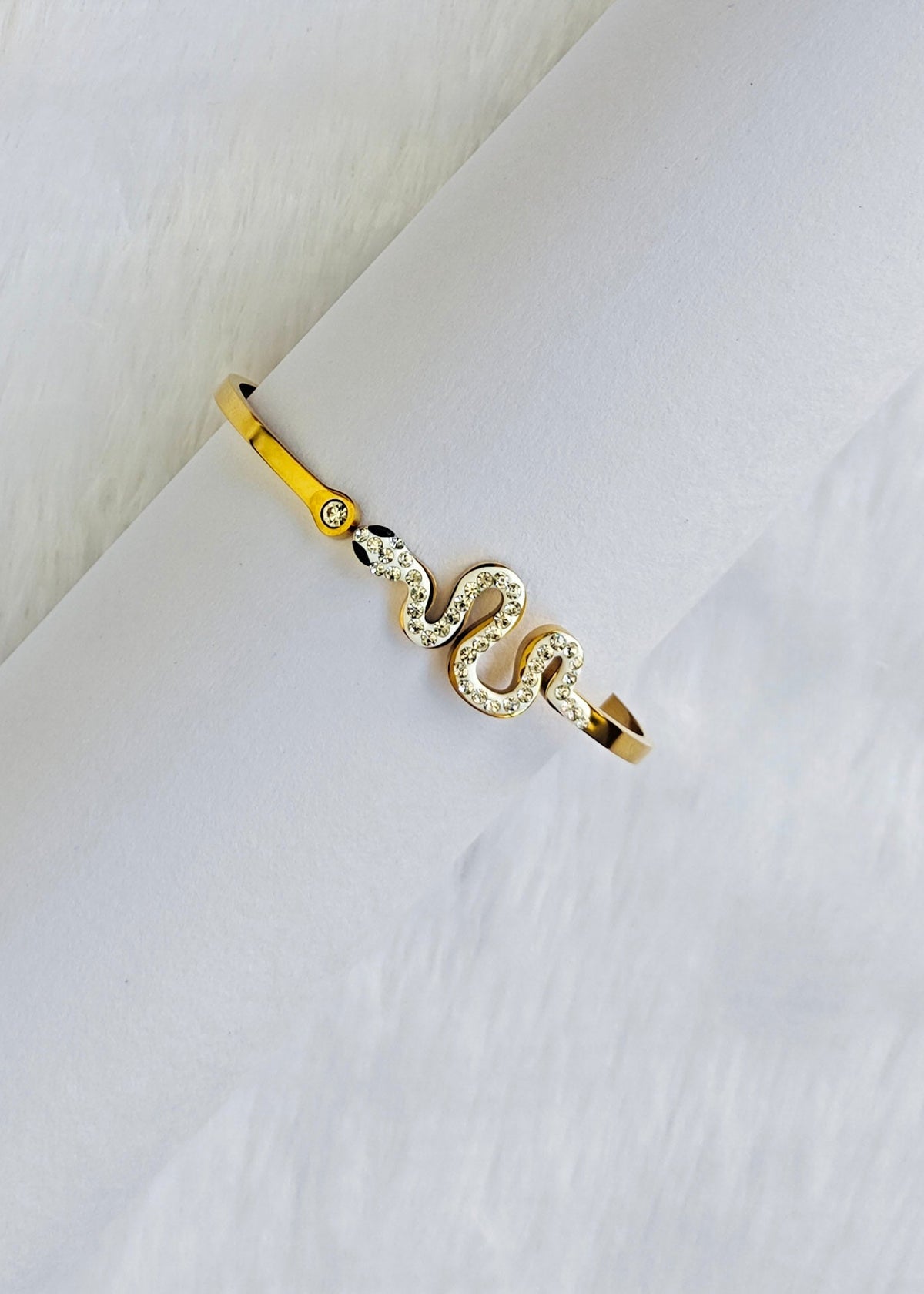A gold snake ring with clear crystal accents, displayed on a white textured surface.