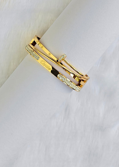 A gold bangle bracelet with a black inlay and pavé crystal accents, shown resting on a white textured surface.