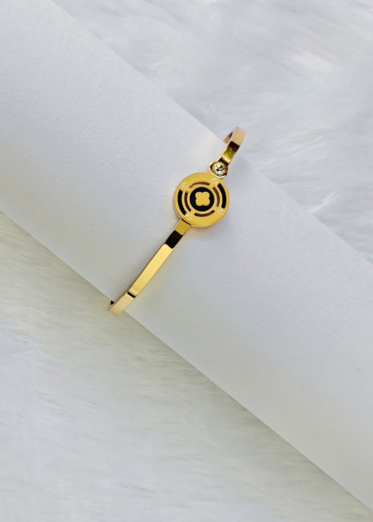 A gold-plated bangle bracelet with a circular medallion charm featuring concentric black rings, displayed on a white textured cylindrical stand against a white faux fur background.