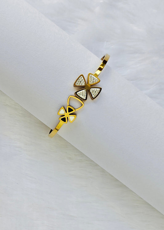 A gold-plated bangle bracelet with three triangular charms, displayed on a white cylindrical stand over a white faux fur background.