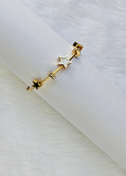 A gold-toned ear cuff bracelet featuring three star charms, including one white star and two gold stars, displayed on a white cylindrical stand over a white faux fur background.
