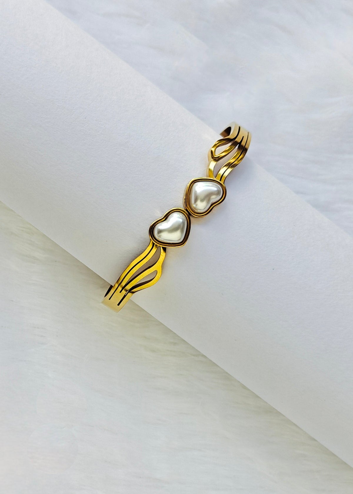 A gold-plated open cuff bracelet with three heart-shaped pearl inlays, displayed on a white textured cylindrical stand.
