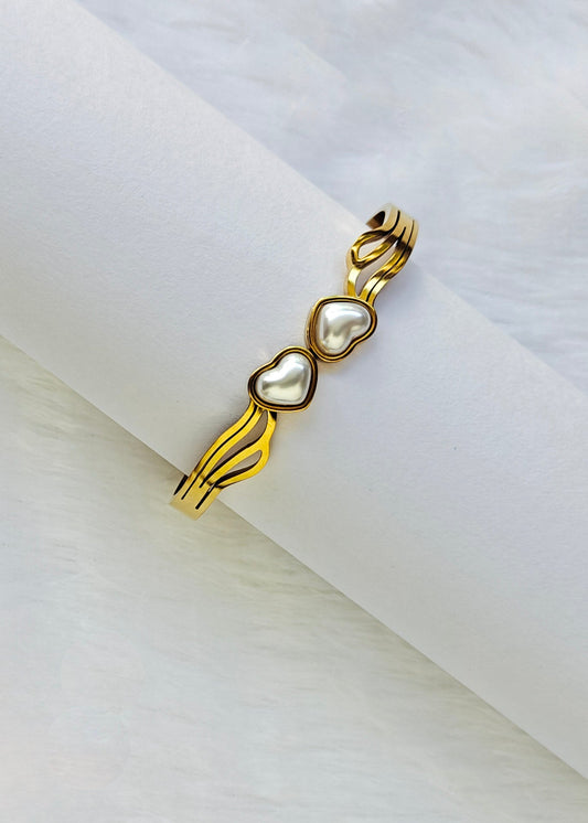 A gold-plated open cuff bracelet with three heart-shaped pearl inlays, displayed on a white textured cylindrical stand.