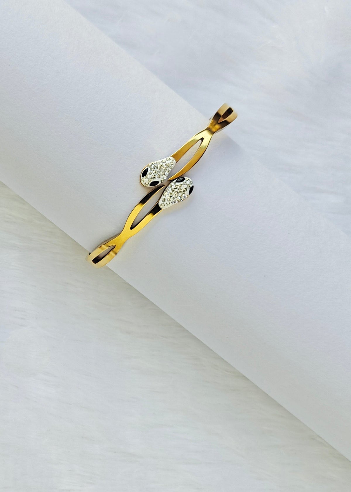 A gold snake cuff ring with clear crystal accents, displayed on a white cylindrical stand against a soft white background.