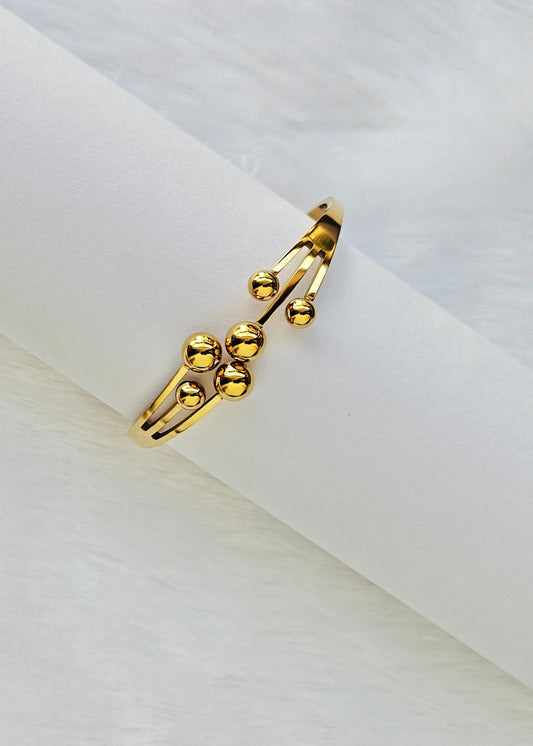 A gold-toned open cuff ring with three small polished bead-like protrusions, displayed on a white cylindrical stand against a soft white background.