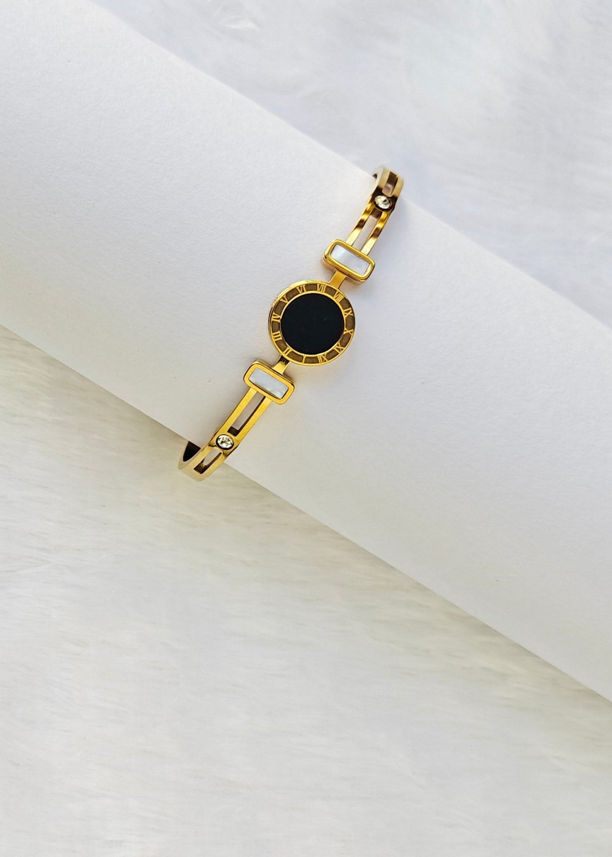 A gold-tone chain bracelet with a central oval black inlay, displayed on a white cylindrical stand against a soft white background.