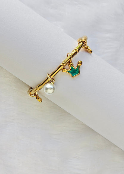 A gold-tone ring with a crown charm, a small green crown accent, and a dangling white pearl, displayed on a white cylindrical ring stand against a soft white background.