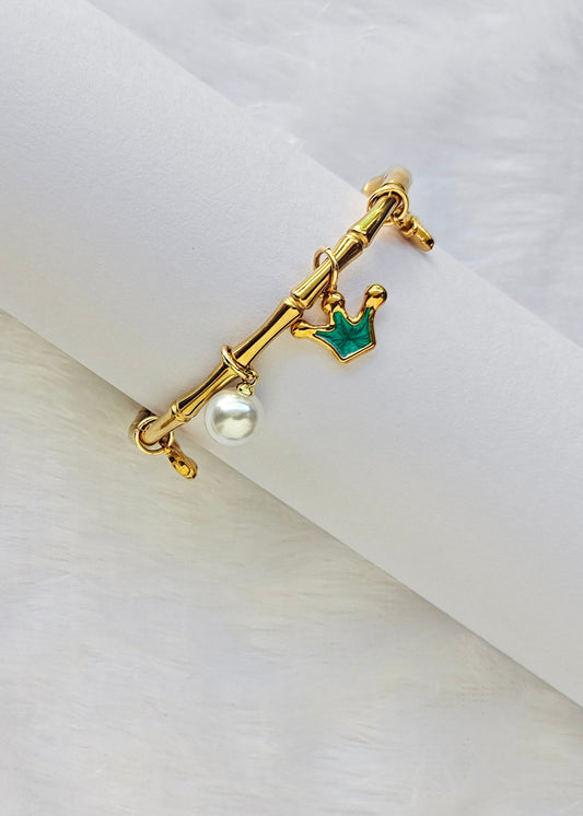 A gold-tone ring with a crown charm, a small green crown accent, and a dangling white pearl, displayed on a white cylindrical ring stand against a soft white background.