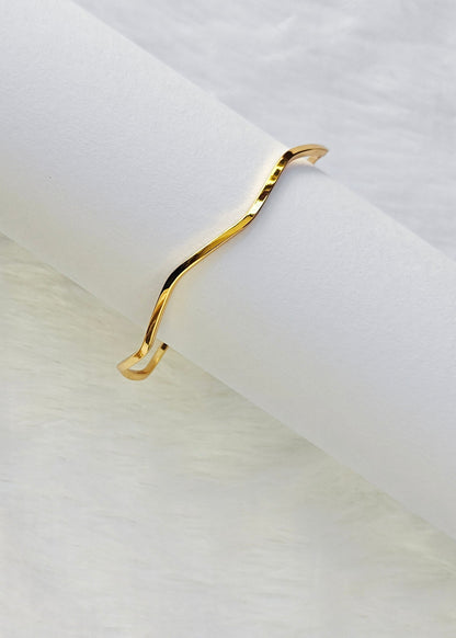 A delicate, wavy open cuff bracelet in polished yellow gold, displayed on a white cylindrical stand against a soft white textured background.
